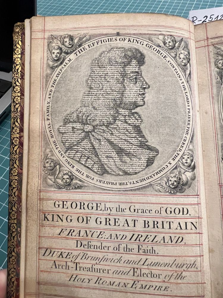 An ornate printed page featuring an engraved portrait of King George I composed entirely of tiny printed text forming his facial features, hair, and clothing. The circular border around the portrait reads: “THE EFFIGIES OF KING GEORGE CONTAINS THE LORD’S PRAYER, THE CREED AND THE X COMMANDMENTS, THE PRAYERS FOR THE KING AND THE ROYAL FAMILY, AND THE XXI PSALM.” Surrounding the border are cherubic faces. Below the portrait, large calligraphic text proclaims King George’s full royal titles in a highly decorative style with ruled red guidelines.