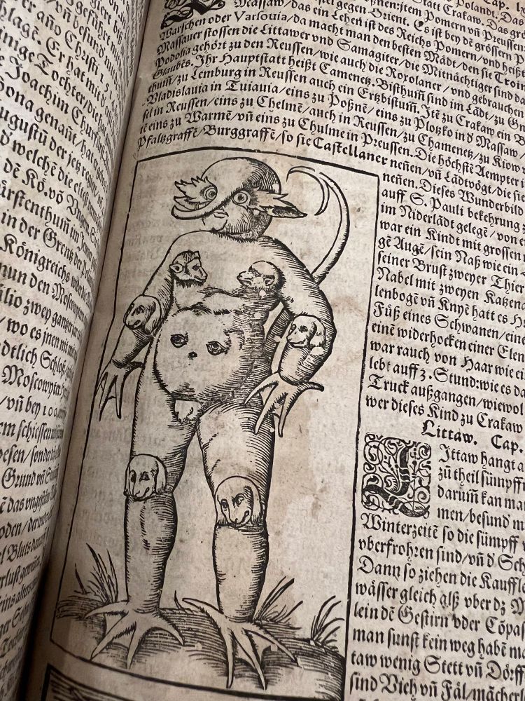 Woodcut illustration from a 16th-century printed book depicting the “Monster of Cracow” — a humanoid figure with multiple animal heads (resembling barking dogs) protruding from its chest, elbows, and knees. The creature has bird-like feet, clawed hands, a distorted face with large eyes, and a long curved tail. The image is surrounded by dense blackletter German text.