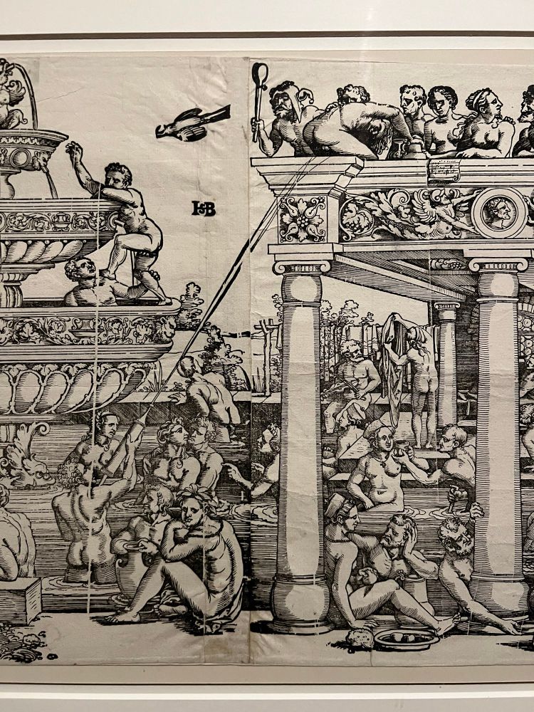 A detailed black-and-white woodcut print showing a crowded public bath scene. Numerous nude and semi-nude figures, both male and female, are seen bathing, washing, socializing, and lounging around an elaborate fountain and classical-style architectural structure with columns. Some figures are scrubbing themselves or each other, while others are engaged in conversation or relaxation.