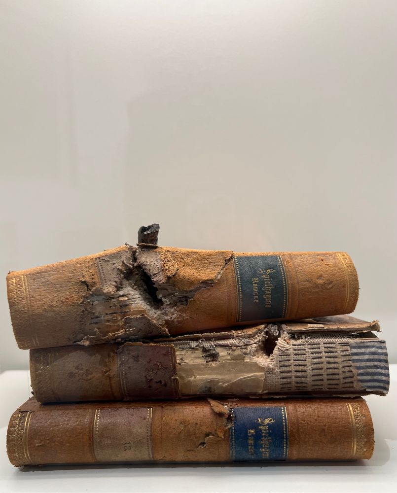 A stack of three old books with visibly damaged spines and covers, pierced by a bullet. The leather bindings are torn, and some pages are shredded and charred.