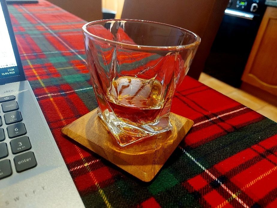 A chunky glass tumbler holding a shot of rum. It stands on a wooden coaster on a red and green tartan check tablecloth. The edge of my writing laptop is visible to the left.