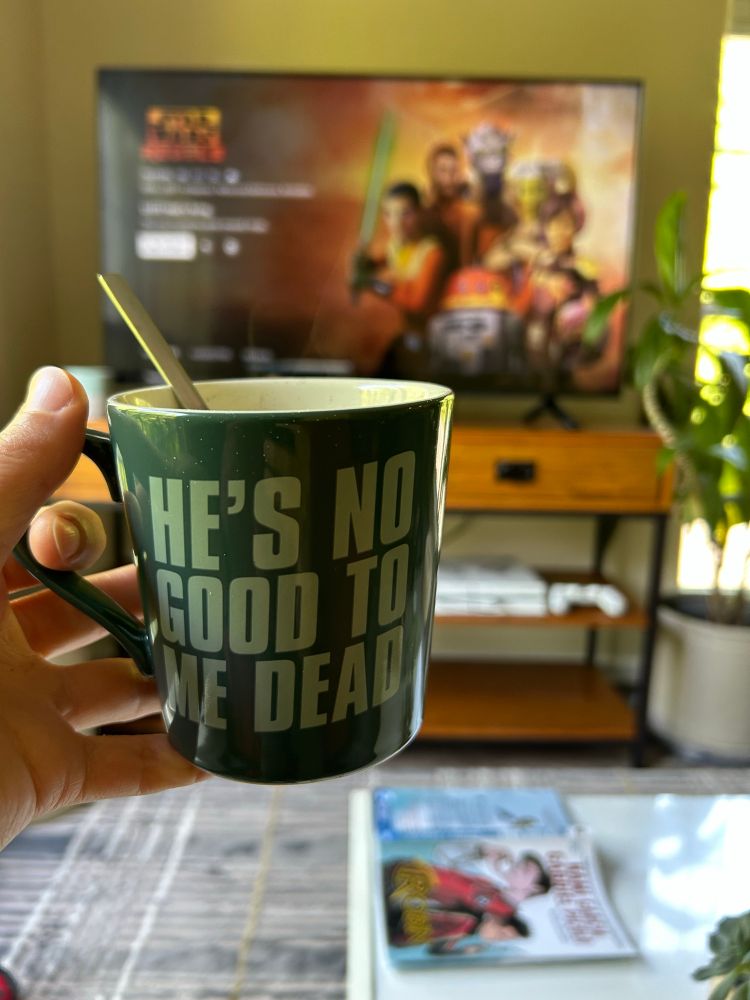 A mug with the Boba Fett’s “He’s no good to me dead” quote is shown with a spoon sticking out of it. In the background is a TV with the Disney+ Star Wars Rebels page pulled up.