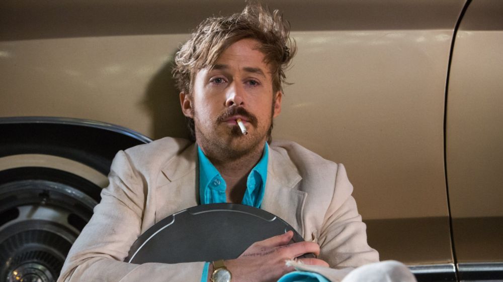 Ryan Gosling in THE NICE GUYS, smoking, dishevelled, clutching a movie canister as he shelters from bullets behind a parked car. 