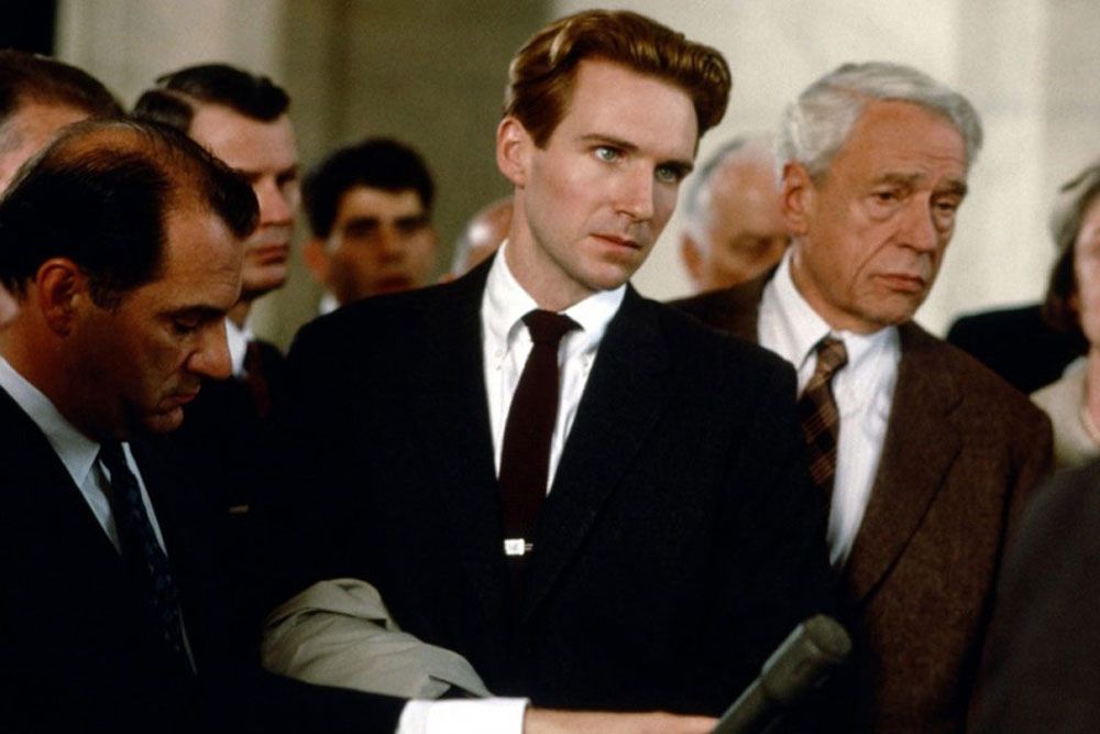 Ralph Fiennes and Paul Scofield in QUIZ SHOW, both in dark suits, looking harrowed as they are hounded by reporters outside a Washington hearing. 