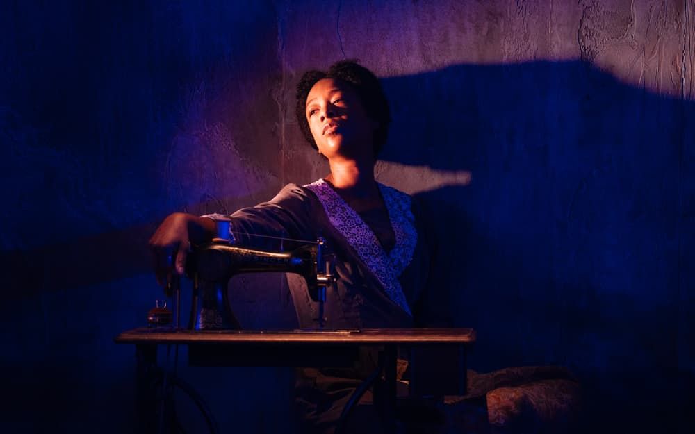 Samira Wiley in Intimate Apparel: a young Black woman sits at a sewing machine in 1905, her face brightly lit, the stage in rich purple shadow. 