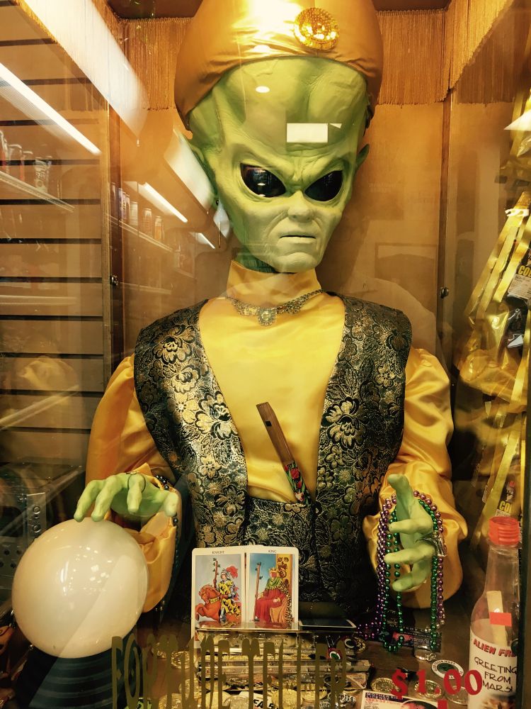 A fortune-telling alien animatronic displayed in a glass booth. The green-skinned alien, dressed in a gold turban, yellow satin shirt, and ornate vest, sits behind a crystal ball with tarot cards, beads, and trinkets on the table. The setting is warmly lit, with reflections of a shop interior visible in the glass.
