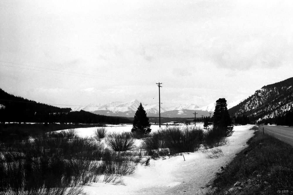 Schwarzweißfoto, aufgenommen in einem Tal in Colorado im Winter mit vereistem Schnee zwischen Büschen und Bäumen im Vordergrund und Blick auf die Kette der Rocky Mountains im Hintergrund. In Bildmitte ein Mast.
