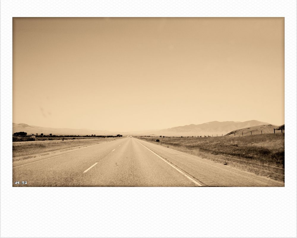 Monochromfoto in Sepia, aufgenommen durch die Windschutzscheibe eines fahrenden Autos irgendwo auf der Autobahn in Montana, USA. Minimalistisches Bild, das die Weite der Landschaft und den riesigen Himmel zeigt (Big Sky ist der Spitzname von Montana).