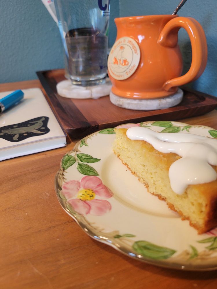 Photo of a slice of lemon cake with cream on top on a floral plate, with an orange mug, glass of water, and notebook in the background