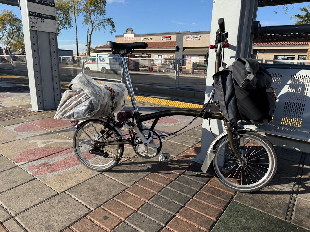 My Brompton bike waiting at the 7th/Camelback Valley MetroRail station. There’s a clear bag lashed to the rear rack and a grey pack attached in front. 