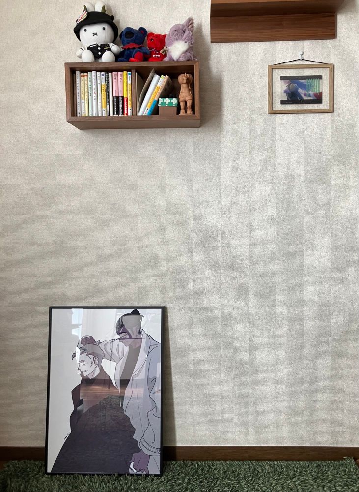 白い壁の前に黒いフレームに入った🐉🔥のポスターが置かれている。床には緑のラグ、壁の上部には本が入った壁掛けの本棚がある。