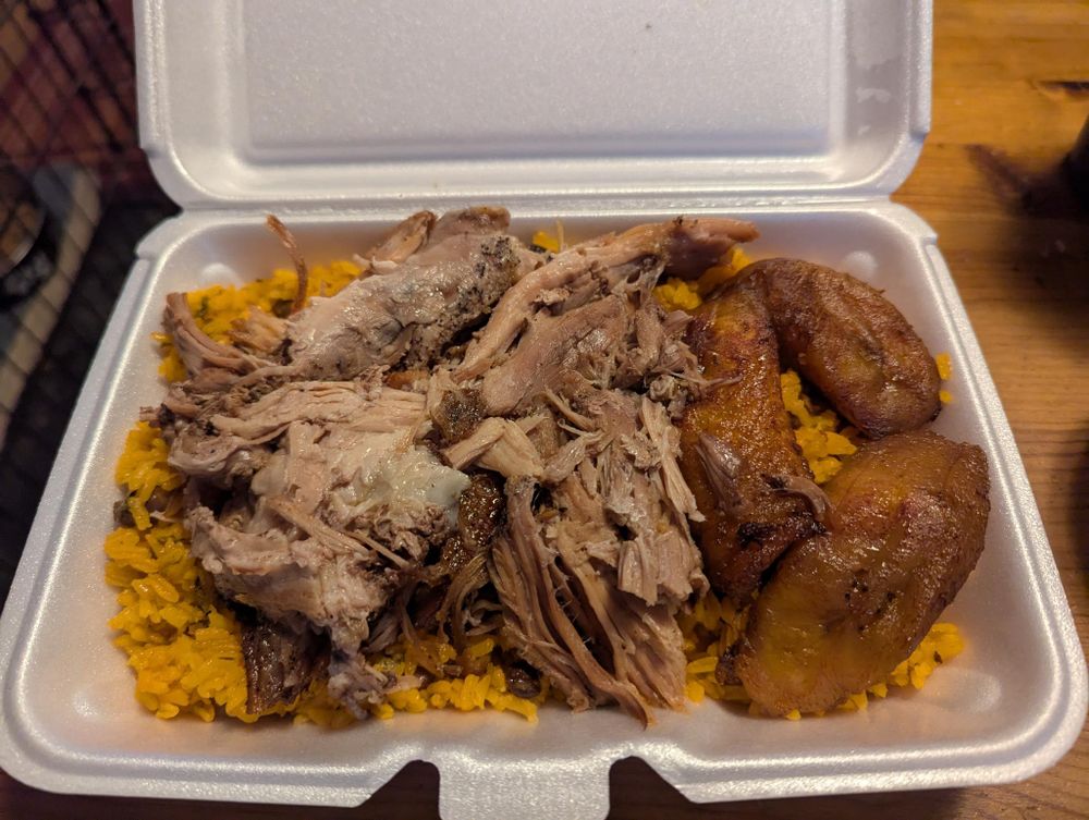 A open styrofoam container with food in it. Sitting on a bed of yellow rice is a mound of shredded, delicious pork, and three fried plantains.