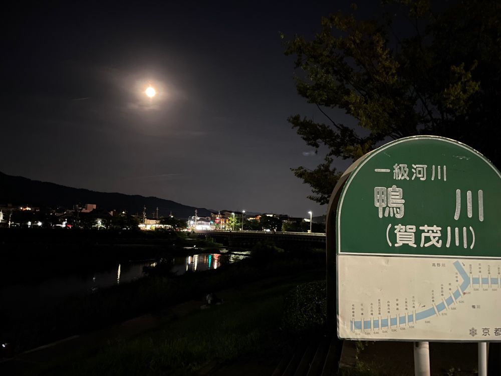 夜、空に明るい月、手前に「一級河川 鴨川（賀茂川）」の看板。奥には鴨川に街の明かりが少し反射し、木や山がある。