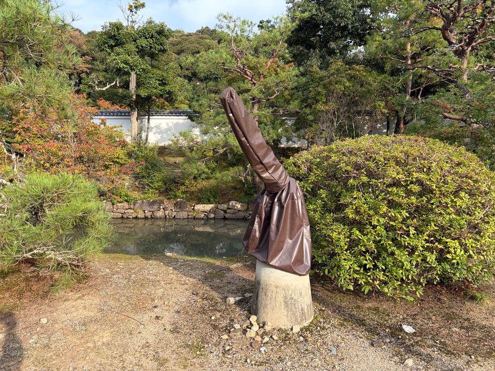 コンクリの土台に、斜め上に伸びた棒状の物体、茶色いビニールが被せられている。背後に丸っこく刈られた木や松など、その向こうに池。奥には紅葉しかけた木、白い壁。雲の多い空。