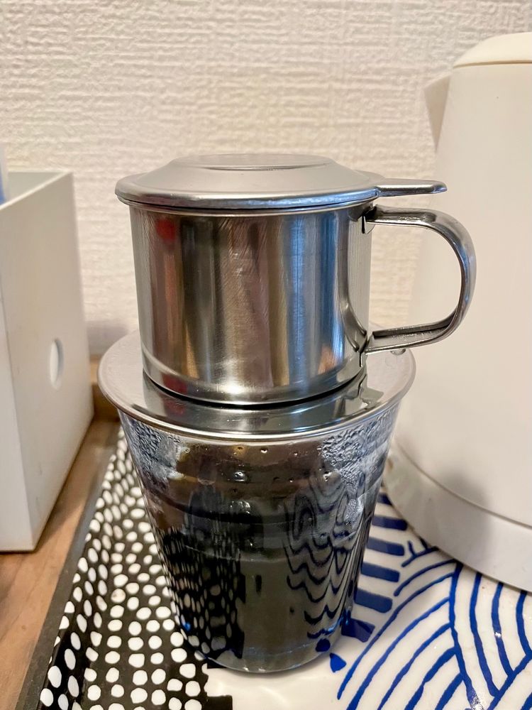 コーヒー抽出中の写真。金属製のフィルター（フィン）がガラスのコップの上に乗っている。フィンはマグカップのような取手のついた、シルクハットみたいな形をしていて、上に蓋がある。コップにはコーヒーが半分ぐらい入っている。右に白い湯沸かしケトル、青波と白黒ドットの柄のお盆の上。左に白い箱、木製の棚の上。