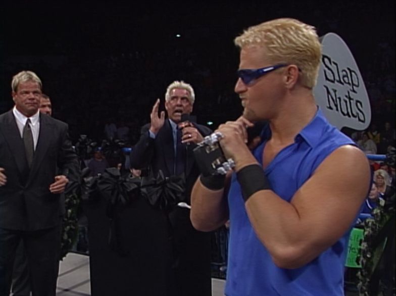 jeff jarrett in a blue sleeveless button up with blue sunglasses holding his patented guitar while ric flair eulogizes the end of goldberg's career.