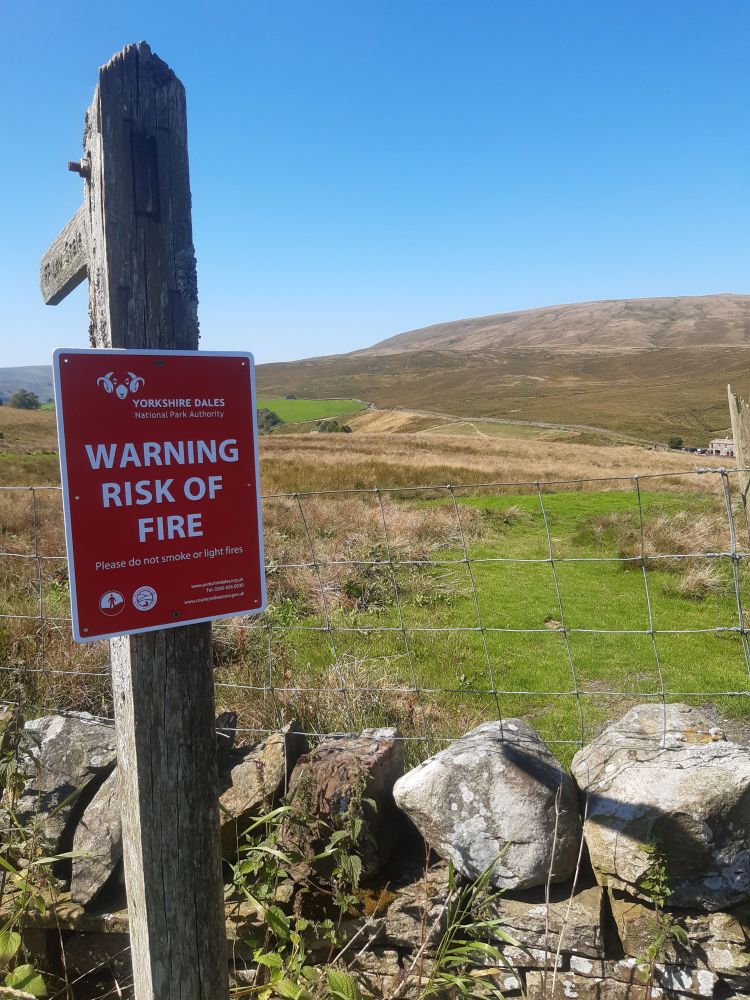 A red warning sign is attached to a wooden post next to a wire fence in a rural area. The sign reads 'YORKSHIRE DALES NATIONAL PARK AUTHORITY WARNING RISK OF FIRE Please do not smoke or light fires.' The background shows an open field with green grass, rocks, and hills under a clear blue sky
