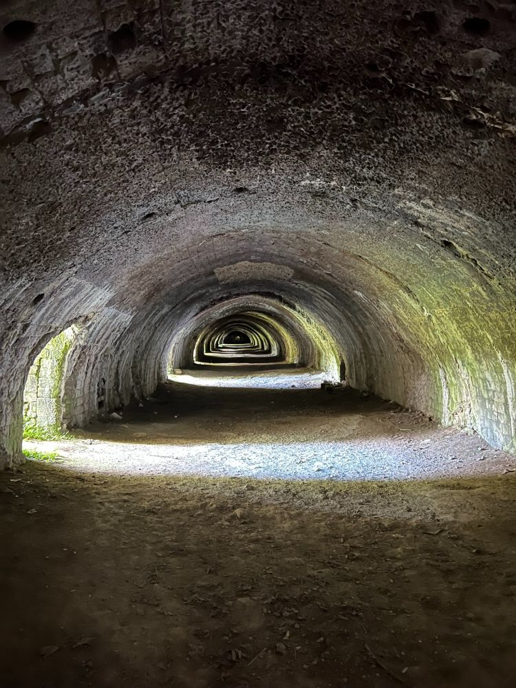 A long, arched tunnel with a dirt floor and weathered stone walls, illuminated by natural light from the entrance. The light gradually fades into darkness deeper into the tunnel. No people or objects are visible