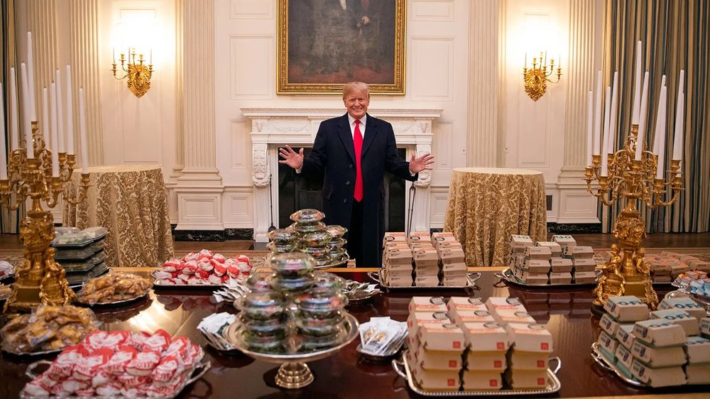 Trump proudly displays a feast of probably-cold McDonald’s amongst gaudy gold candelabras in the White House during his first term as President, with his hands splayed open and a shit-eating grin on his face.