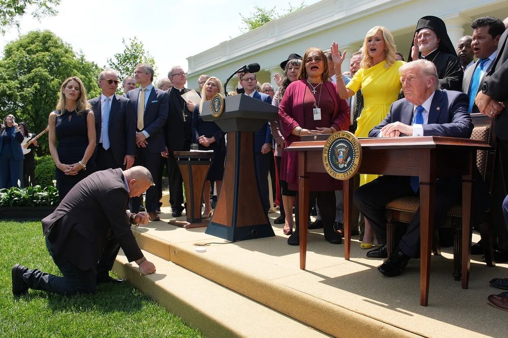 Trump at a desk surrounded by religious and political leaders who look a whole lot like they're worshipping him, including one who's literally taking a knee in front of Trump like he's bowing to Caesar.