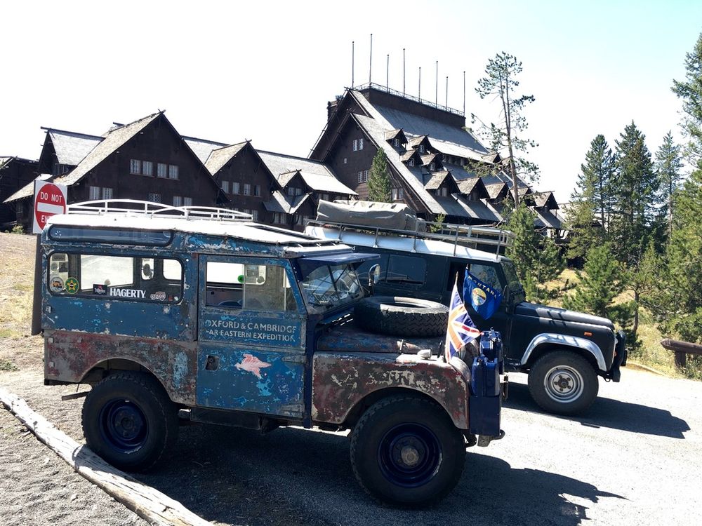 A faded blue and corroded Series 1 Land Rover 86-Inch parked in front of the Old Faithful Inn, Yellowstone National Park. A black and silver Defender 110 is parked behind it.