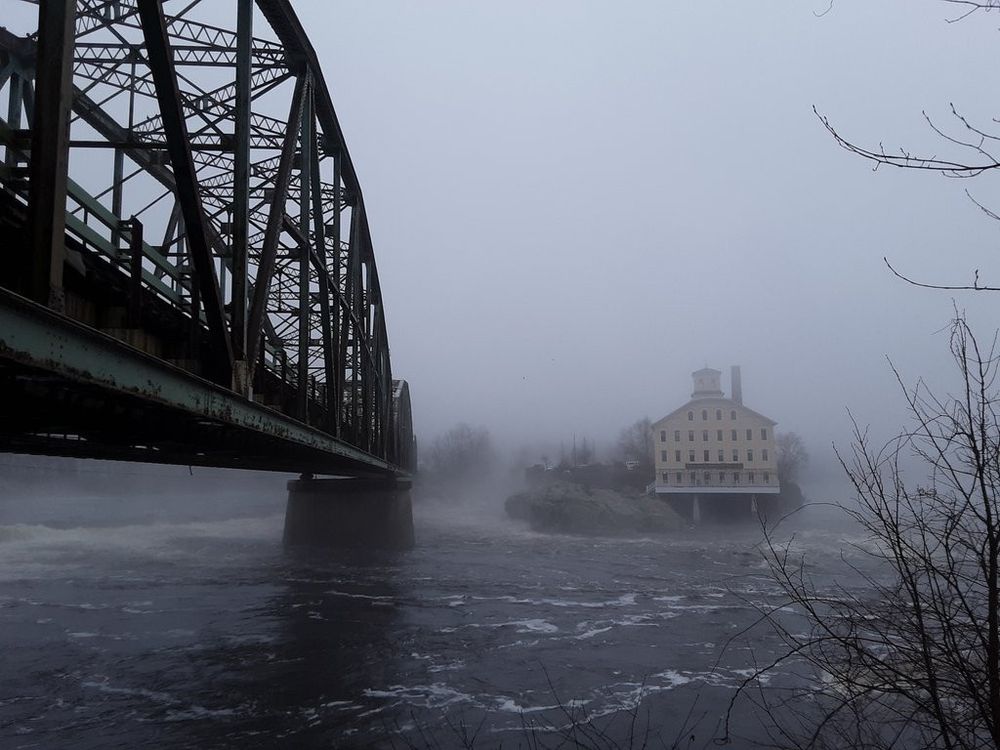 a photo of a rusty green bridge and a distant building over a river, caked in fog