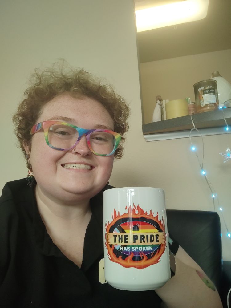 A genderqueer person with rainbow glasses smiles facing camera while holding their mug of tea with The Pride Has Spoken logo facing outward and centered.