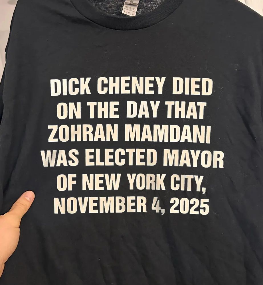 A black T-shirt with white text (Impact Font) that reads: "DICK CHENEY DIED ON THE DAY THAT ZOHRAN MOMDANI WAS ELECTED MAYOR OF NEW YORK CITY NOVEMBER 4, 2025"