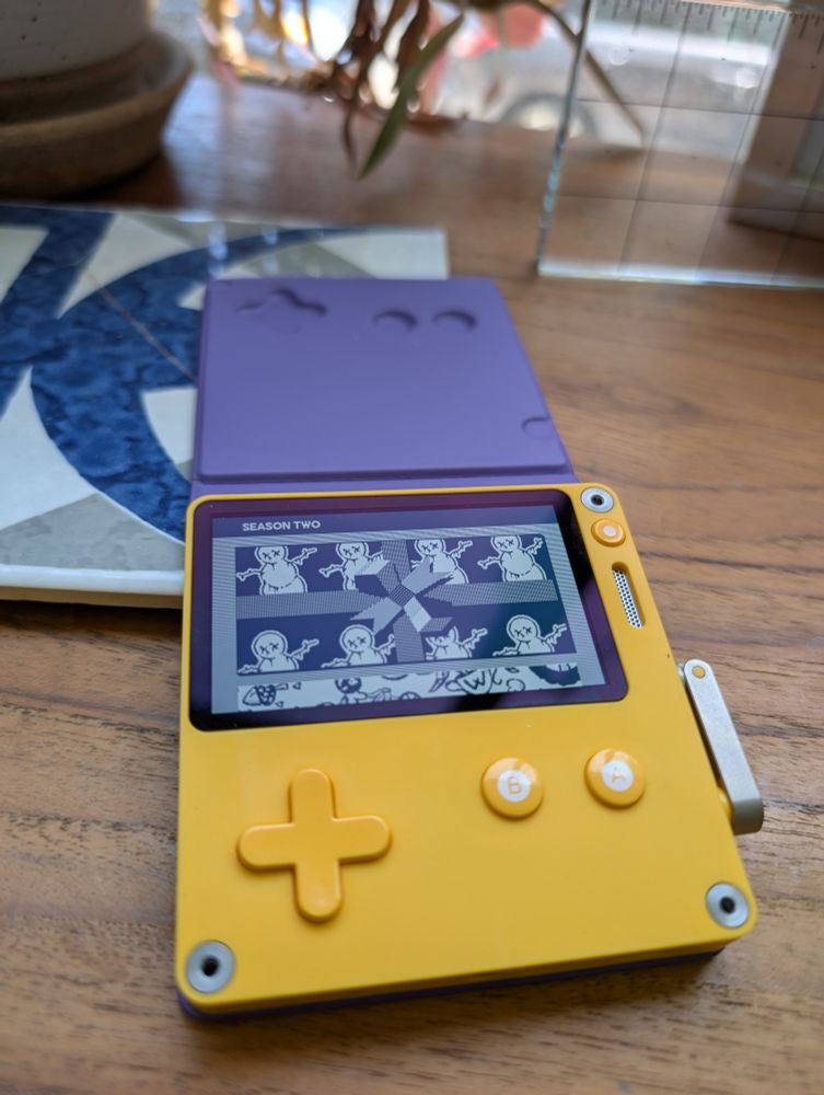 A yellow gaming device with a crank on a wood table.  It has a LCD screen with an image of a present and what look to be snowmen.  There is a plant in the background. 