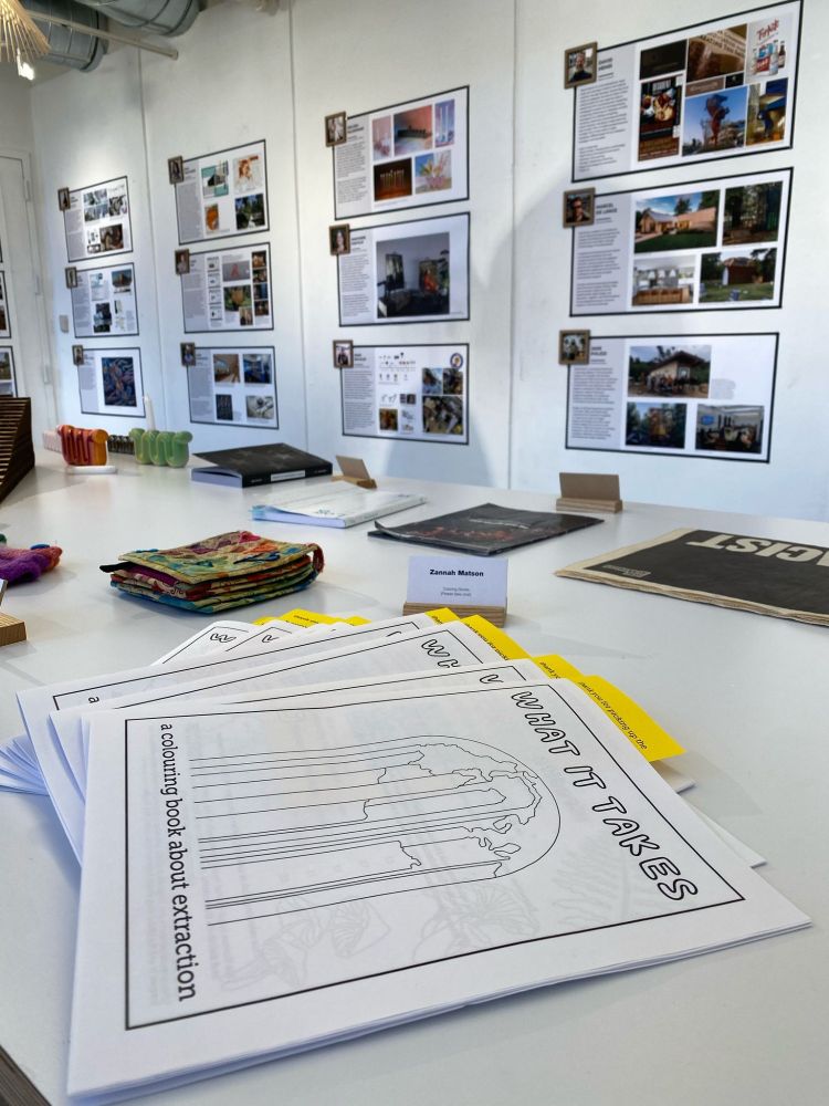 Pile of white colouring books on a table in a white-walled gallery space with various other media in the background