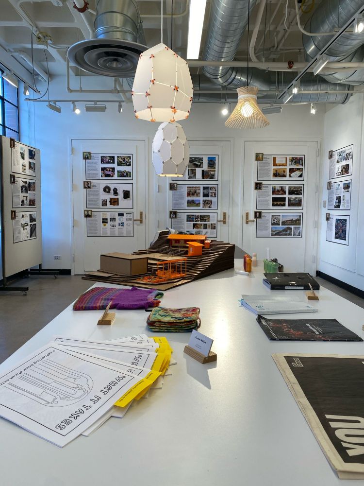 White table with various media in a white-walled gallery space with posters on the walls and white and beige light fixtures hanging 