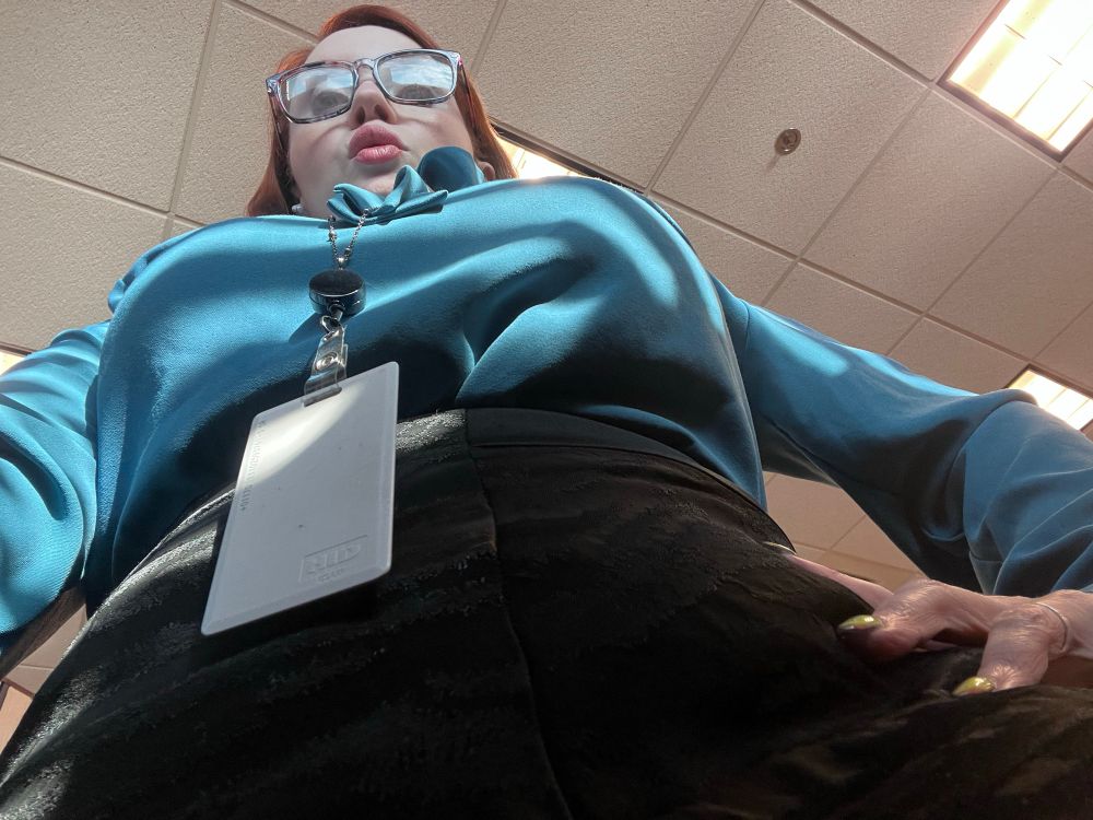 A view of a white redhead wearing glasses, a teal blouse, black skirt and lanyard. 