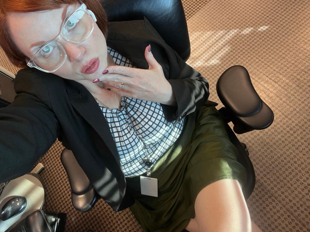 White redhead in an office chair wearing clear glasses, a black blazer, white grid patterned blouse and a green satin skirt. 