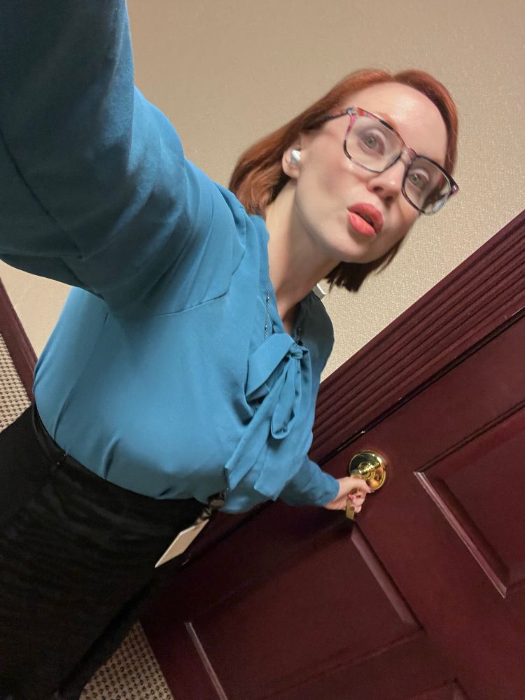 A view of a white redhead wearing glasses, a teal blouse, black skirt and lanyard. She’s opening a door. 