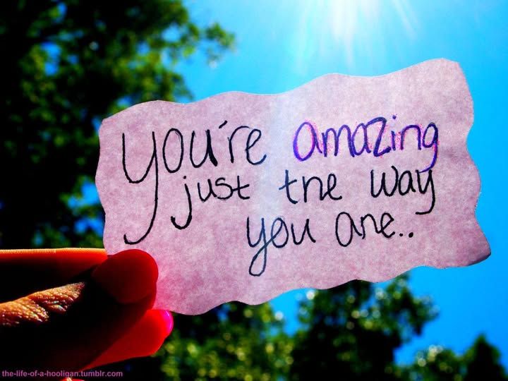 image is of a small scrap of paper, being held up in the sunlight outdoors. In the background, blue skies and green trees can be seen. On the scarp of paper, there is a hand-written message in ink: "You're amazing just the way you are."