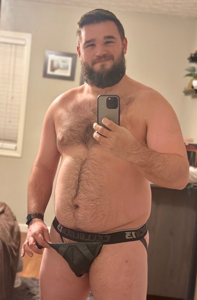 A man wearing nothing but a jockstrap takes a selfie in a mirror. One hand is holding the front flap of the jockstrap open. 