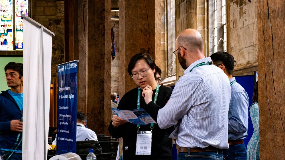 Two people talk while looking at some leaflets. There are other people and banners in the background.