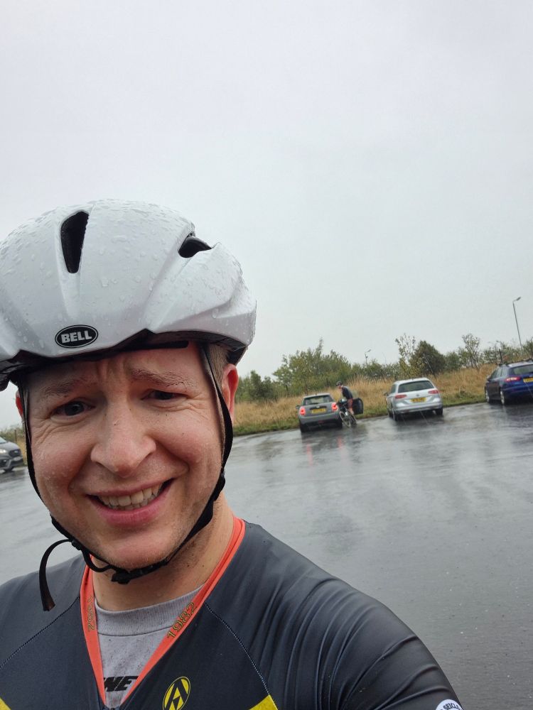 A man wearing a black cycling jersey and white helmet, standing in a car park in the pouring rain.