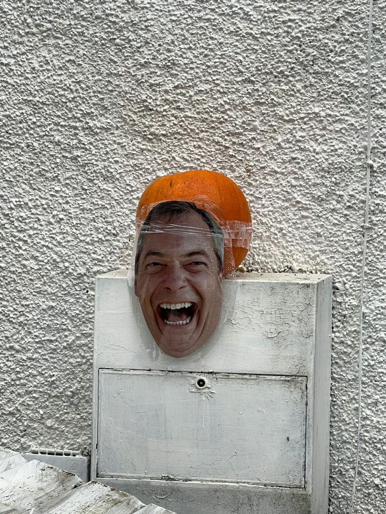 A paper mask of Nigel Farage's face taped to a pumpkin, sitting on a white box outside a white pebble-dashed house