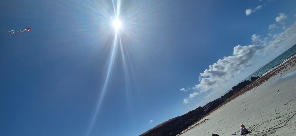 A small red kite flying in a blue sky on a sandy beach, with low dark cliffs on the left, and the sun on the right
