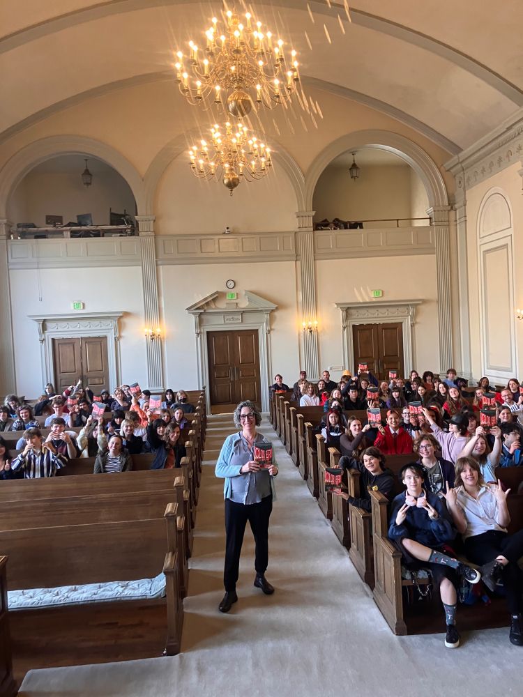 A photo of me holding Closer inside a huge room of Northwest Academy high school students holding their copies of Closer. A couple chandeliers hang overhead. 