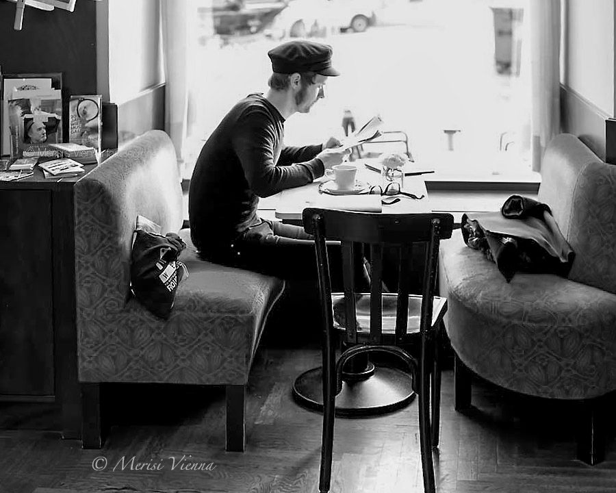A young man with a Breton cap sitting on an upholstered bench - a coffee table in front of him, with a cup of coffee, reading glasses and a small vase with flowers - and reading.