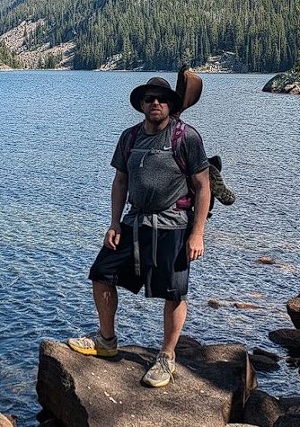 Greg standing in from of a mountain lake in a broad brimmed hat and running shoes.  The legs are bleeding from multiple scratches.