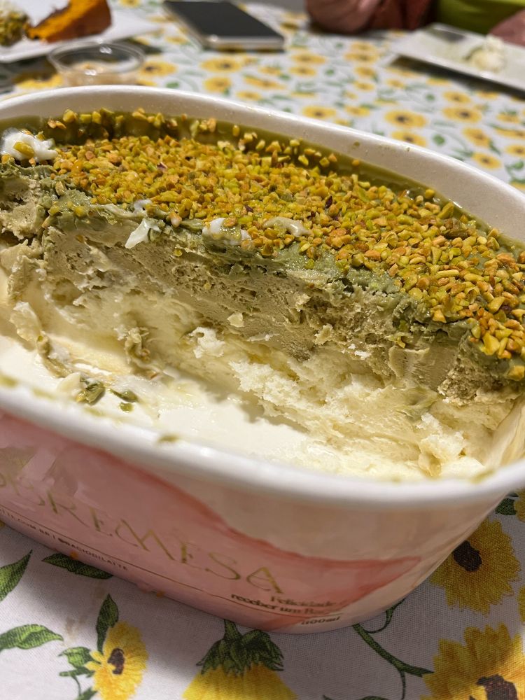 Close numa embalagem oval, em tons de rosa, com metade de um pavê de pistache. Na foto da para ver cada camada do pavê, a de base é cor creme, seguida por uma camada verde claro, uma verde escura e pistaches picados por cima. Ao fundo da imagem, uma toalha de mesa branca com estampa de girassois amarelos.