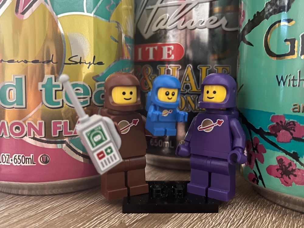 three modern Lego “space” minifigures pose in front of a variety of Arizona Tea cans. one is wearing all brown and carries a baby monitor in one hand, and carries a tiny space baby in a blue suit in the other. the other minifigure is wearing all purple