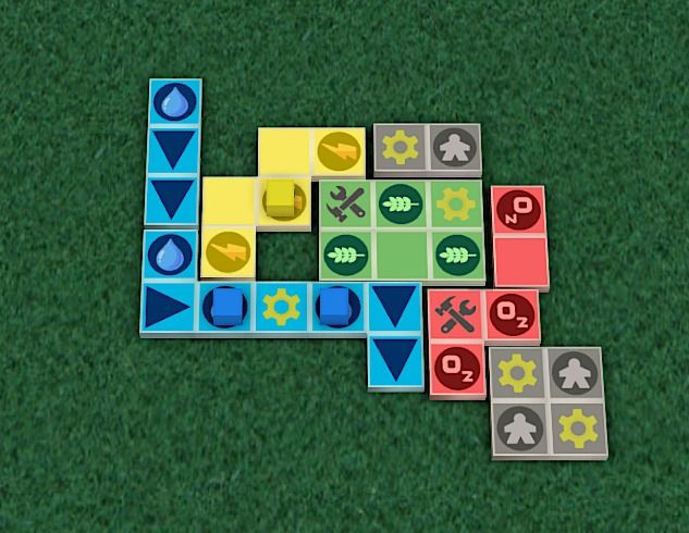 A board game prototype with colorful square tiles arranged on a green surface. The tiles show various symbols, including water drops, gears, tools, wheat, oxygen, meeples, and arrows, in colors like blue, yellow, green, gray, and red.