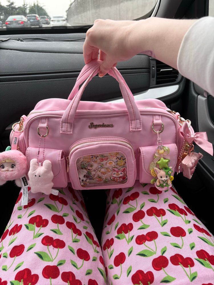 A wide pink bag decorated with many cute keychains and pins is being held in the lap of a person wearing pink cherry print overalls 