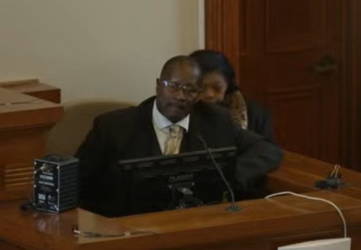 Man in a suit with a woman behind him both sitting in a witness stand in a courtroom