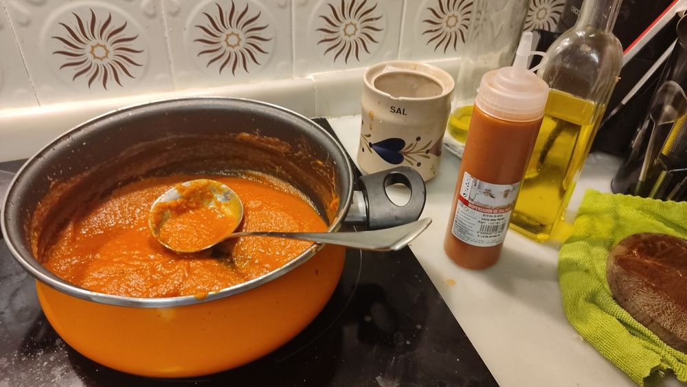 Foto del banco de una cocina.
En la vitrocerámica hay una olla con salsa brava de un color naranja rojizo, sobre el mármol se ve un biberón para salsas lleno, un salero de cocina, dos aceiteras de aceite de oliva, un trapo y una tabla de cortar de madera muy pequeña.