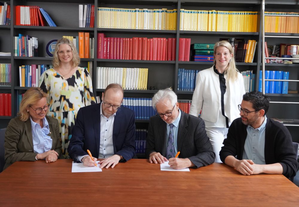Man sieht insgesamt sechs Personen des IDS und der Marie-Curie-Realschule bei der Unterzeichnung der Kooperationsvereinbarung. Und zwar: Gabriele Herrmann-Krotz, Heike Chan Hin und Henning Lobin vom IDS (linke Seite) und Hendrik Tzschaschel, Natascha Werning und Mehmet Bayrakli von der Marie-Curie-Realschule (rechte Seite). In der Mitte unterzeichnen Herr Lobin und Herr Tzschaschel die Kooperationsvereinbarung am Tisch sitzend, die anderen Personen stehen neben ihnen. Hinter den Personen ist eine Bücherwand.